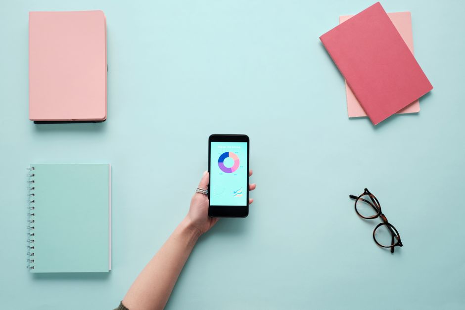 Minimalist flat lay with a smartphone displaying charts, surrounded by notebooks and glasses on a blue background.